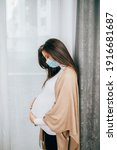 Small photo of Young pregnant woman is standing by window with face protective mask. She is worried about childbirth because od Covid-19 pandemic.