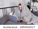 Young attractive caucasian man sitting on a bed wrapped with blanked at home working using laptop with cup of hot drink. Guy browsing Internet for flu, influenza, cold and covid-19 symptoms