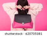 Woman in a pink coat holding handbag with two hands behind the back on a pink background