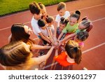 Small photo of A large group of children, boys and girls, stand together in a circle and fold their hands, tuning up and raising team spirit before the game at the stadium during sunset. A healthy lifestyle.
