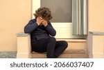 Small photo of emotional frailty and crying for a school-age child - violence in the family and child abuse - 7-year-old child sitting on the ground outside the home is in despair