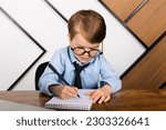 Small photo of Little boy in round eyeglasses, blue shirt and tie sitting at the desk in the office with a notepad. Smart little kid, Child prodigy.