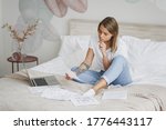Concerned young woman girl in white t-shirt sitting in bed with white sheet pillow blanket spending time in bedroom at home. Remote work study lifestyle concept. Working with laptop papers documents