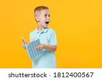 Small photo of Little fun male fair-haired brown-eyed kid boy 5-6 years old wearing stylish blue turquoise t-shirt polo holding in hands reading book isolated on yellow color wall background, child studio portrait