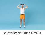 Full length portrait of shocked strong young fitness man with skinny body sportsman in headband shirt shorts showing biceps muscles isolated on blue background. Workout gym sport motivation concept