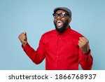Excited amazed young bearded african american man 20s wearing casual red shirt cap eyeglasses standing doing winner gesture clenching fists isolated on pastel blue color background studio portrait