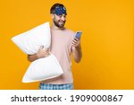 Smiling young man in pajamas home wear sleep mask hold pillow using mobile cell phone typing sms message while resting at home isolated on yellow background studio portrait. Relax good mood concept