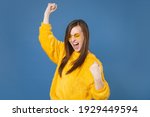 Happy joyful screaming young brunette woman 20s wearing yellow fur sweater eyeglasses posing clenching fists doing winner gesture keeping eyes closed isolated on blue color background studio portrait