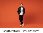 Full size length smiling cheerful young bearded man 20s wearing basic white t-shirt black leather jacket standing holding hands in pockets looking camera isolated on orange background studio portrait
