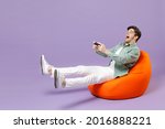 Full length man in casual mint shirt white t-shirt sitting in orange bean bag chair hold takeaway bucket eat popcorn watch movie film play pc game with joystick console isolated on purple background.