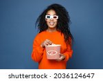 Cheerful young african american woman in casual basic bright orange sweatshirt 3d glasses standing watching movie film, holding bucket of popcorn isolated on blue color background studio portrait