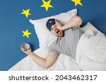 Top view young shocked latecomer man in pajamas jam sleep mask rest at home lies wrap covered under blanket hold face slept late isolated on dark blue sky background Bad mood night bedtime concept.