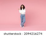 Full length young smiling cute african american woman 20s curly hair wear white casual knitted sweater jeans looking camera hold hands crossed folded isolated on pastel pink background studio portrait