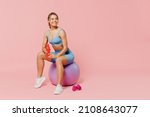 Full length young athletic fitness trainer instructor woman wear blue tracksuit spend time in home gym sit on fitball drink water isolated on pastel plain light pink background Workout sport concept