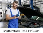 Small photo of Young professional technician car mechanic man 20s in blue overalls white t-shirt use hold mobile cell phone browsing fix problem with raised hood bonnet work in vehicle repair shop workshop indoor.