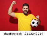Small photo of Excited happy young bearded man football fan in yellow t-shirt cheer up support favorite team hold soccer ball celebrate clenching fists say yes isolated on plain dark red background studio portrait