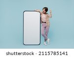 Small photo of Full body young smiling happy woman 20s in casual brown shirt stand near big mobile cell phone with blank screen workspace area do winner gesture isolated on pastel plain light blue color background.