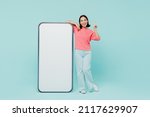 Full body young woman of Asian ethnicity in pink sweater stand near big mobile cell phone with blank screen workspace area point finger up with new idea isolated on pastel plain light blue background