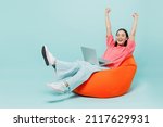Full body young happy woman of Asian ethnicity 20s in pink sweater sit in bag chair use work on laptop pc computer with outstretched hands finish job isolated on pastel plain light blue background.