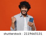 Small photo of Young bearded Indian man 20s years old wears blue shirt hold in hand use mobile cell phone doing winner gesture celebrate clenching fists say yes isolated on plain orange background studio portrait