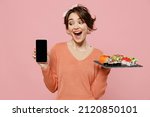 Small photo of Young surprised woman in sweater hold makizushi sushi roll served on black plate traditional japanese food use mobile cell phone blank screen workspace area isolated on plain pastel pink background.