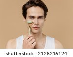Small photo of Young man he 20s perfect skin in undershirt doing face massage with jade facial roller natural quartz stones isolated on pastel light beige background. Skin care healthcare cosmetic procedures concept