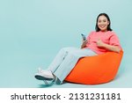 Full body young smiling happy woman of Asian ethnicity 20s wearing pink sweater sit in bag chair hold in hand use mobile cell phone show thumb up gesture isolated on pastel plain light blue background