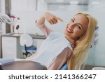Small photo of Young smiling happy fun woman 20s covered by napkin point index finger on healthy toothy smile sit at dental office chair indoor light cabinet wait for stomatologist Healthcare oral enamel treatment.