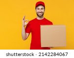 Professional delivery guy employee man 20s in red cap T-shirt uniform workwear work as dealer courier hold face with cardboard box show ok gesture isolated on plain yellow background. Service concept