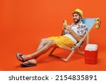 Small photo of Young fun happy tourist man in beach shirt hat lie on deckchair near fridge hold use mobile cell phone isolated on plain orange background studio portrait. Summer vacation sea rest sun tan concept.