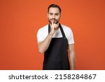Small photo of Young secret man barista bartender barman employee in black apron white t-shirt work in coffee shop say hush be quiet finger on lips shhh gesture isolated on orange background Small business startup