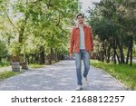 Full length young man 20s he wearing orange jacket blue t-shirt walking look camera rest relax in spring green city park go down alley sunshine lawn outdoors on nature. Urban lifestyle leisure concept