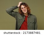 Small photo of Confused disconcerted unnerved aggrieved upset young brunet curly man 20s wears khaki shirt glasses looking camera put hands on head scratching isolated on plain olive green background studio portrait