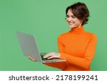 Young smiling copywriter happy fun woman 20s wear orange turtleneck hold use work on laptop pc computer isolated on plain pastel light green color background studio portrait. People lifestyle concept
