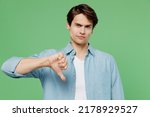 Small photo of Distempered unnerved aggrieved upset disconcerted young brunet man 20s years old wears blue shirt showing thumb down dislike gesture looking camera isolated on plain green background studio portrait