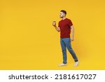 Full length side view smiling young man 20s in red t-shirt casual clothes hold takeaway delivery craft paper brown cup coffee to go walk isolated on plain yellow color wall background studio portrait.