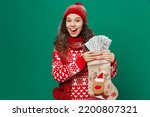 Merry young woman wear red warm cozy knitted sweater hat posing hold Christmas sock fan of cash money in dollar banknotes isolated on plain dark green background. Happy New Year 2023 holiday concept