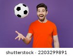 Small photo of Young cheerful cool surprised excited satisfied fun fan man wears orange t-shirt cheer up support football sport team watch tv live stream toss up soccer ball 2024 isolated on plain purple background
