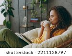Sideways young woman of African American ethnicity wear t-shirt drink coffee read book close eyes sits in armchair stay at home flat rest relax spend free spare time in living room indoor grey wall