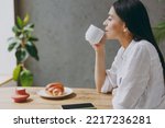 Side view young calm minded latin woman 30s wears white shirt hold cup drink coffee eat croissant sit alone at table in coffee shop cafe restaurant indoors. Freelance mobile office business concept