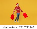 Full body side view young man wear red knitted christmas sweater Santa hat hold shopping paper package bags walk go isolated on plain yellow background. Black Friday New Year 2023 sale buy day concept