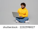 Small photo of Full body happy young IT Indian man 20s he wearing casual yellow hoody sitting using holding working on laptop pc computer isolated on plain grey background studio portrait. People lifestyle portrait