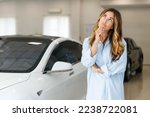 Small photo of Pensive woman 20s customer female buyer client wears blue shirt prop up chin chooses auto wants to buy new automobile in car showroom vehicle salon dealership store motor show indoor. Sales concept.