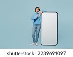 Small photo of Full body amazed young woman wear knitted sweater point index finger big huge blank screen mobile cell phone smartphone with workspace mockup area isolated on plain pastel light blue cyan background