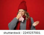 Young sad woman wearing grey sweater scarf hat hold napkin tissue blowing nose sneeze isolated on plain red background studio portrait Healthy lifestyle ill sick disease treatment cold season concept