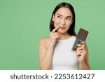 Small photo of Young fun woman wears white clothes hold in hand dark chocolate bar prop up face look aside isolated on plain pastel light green background. Proper nutrition healthy fast food unhealthy choice concept