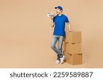Full body smiling delivery guy employee man wears blue cap t-shirt uniform workwear work as dealer courier stand near stack of cardboard boxes scream in megaphone isolated on plain beige background