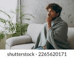 Young sleepy exhausted tired sick ill Indian man wapped in plaid yawn cover mouth with hand sits on grey sofa couch stay at home hotel flat spend free spare time in living room indoor Lounge concept