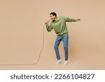 Full body expressive fun young man of African American ethnicity wear green sweatshirt sing song in microphone isolated on plain pastel light beige background studio portrait. People lifestyle concept