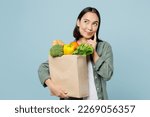 Small photo of Young woman wear casual clothes hold brown paper bag with food products put hand prop up on chin look aside isolated on plain blue background studio portrait. Delivery service from shop or restaurant